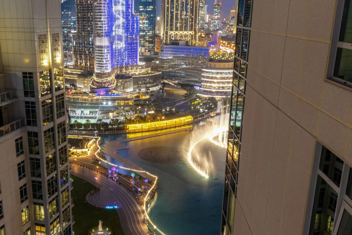 Nighttime cityscape viewed between buildings, with illuminated towers and waterfront roads.