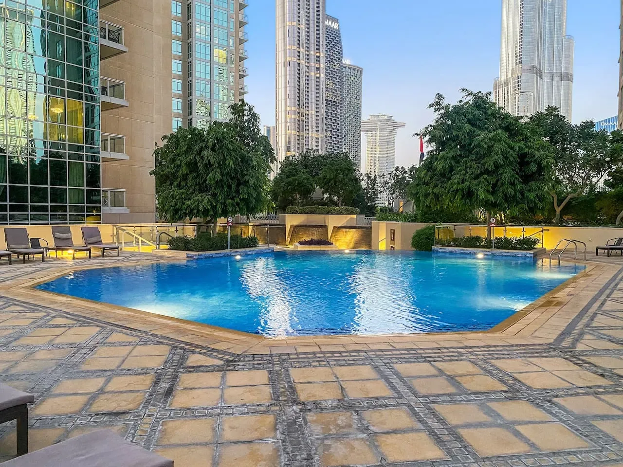 An outdoor swimming pool sits among landscaped grounds and high-rise buildings.