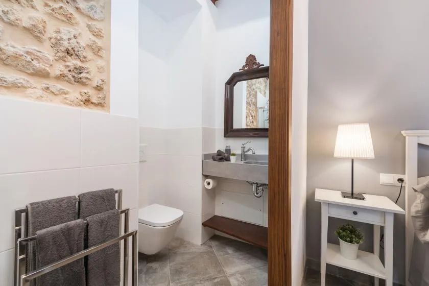 Un baño con lavabo, espejo e inodoro junto a una mesita con una lámpara.