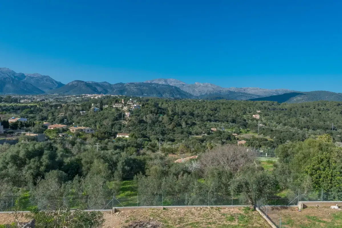 Una vista amplia del paisaje muestra colinas arboladas, casas dispersas y montañas bajo un cielo despejado.