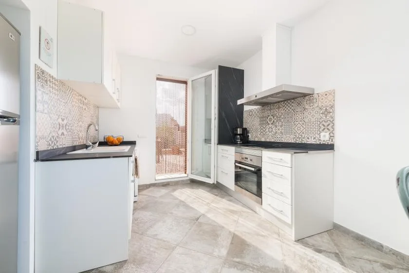 Una cocina luminosa con gabinetes blancos, encimeras oscuras y azulejos estampados.