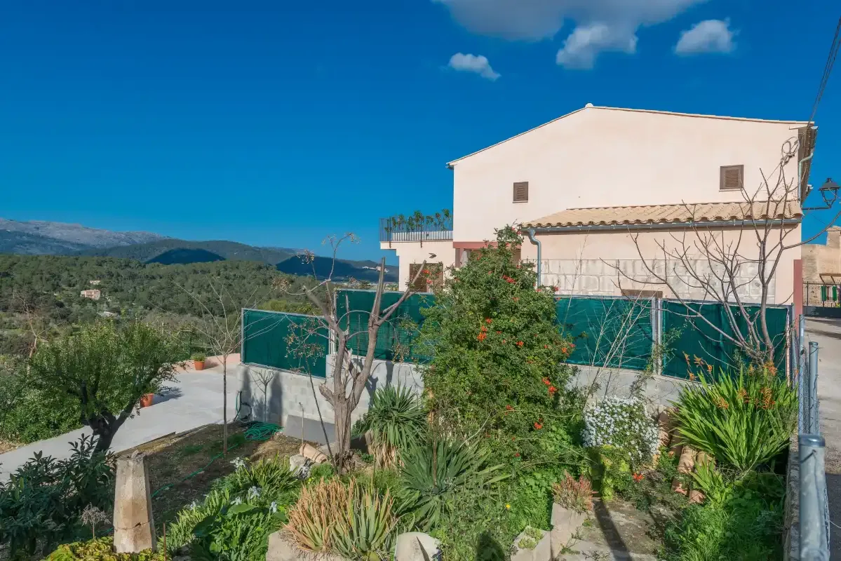 Una casa se alza detrás de un jardín y una valla verde, con colinas a lo lejos.