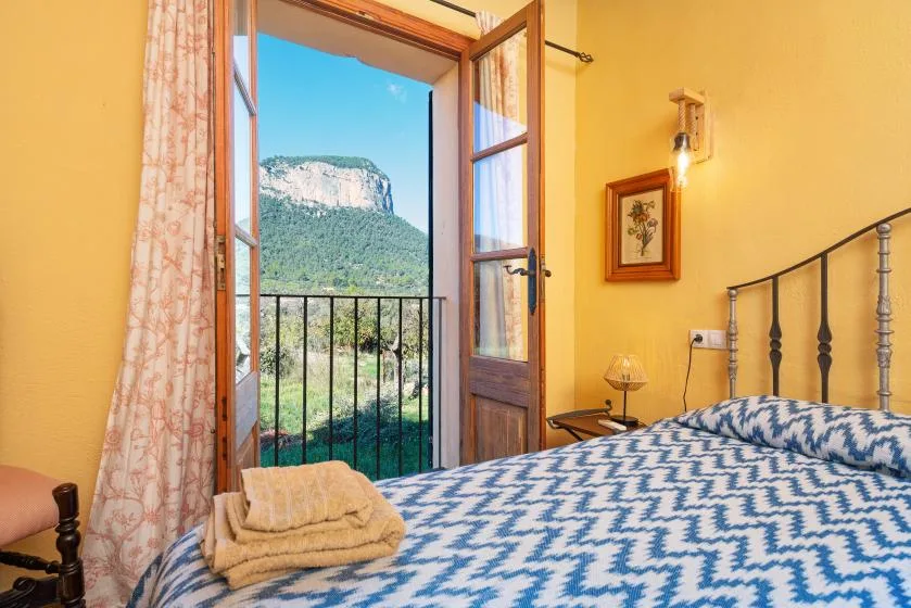 A bedroom with a patterned bed beside an open balcony door overlooking a green hillside.