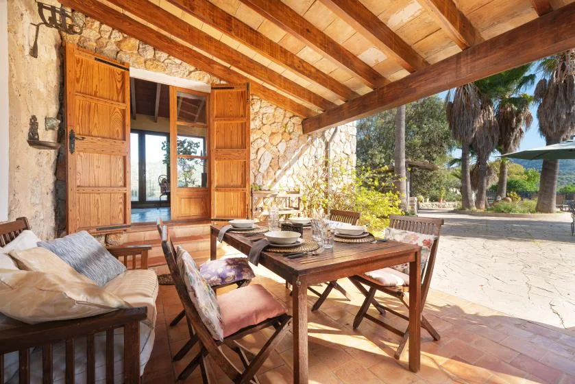 A covered outdoor dining area with a wooden table and seating beside open wooden doors.