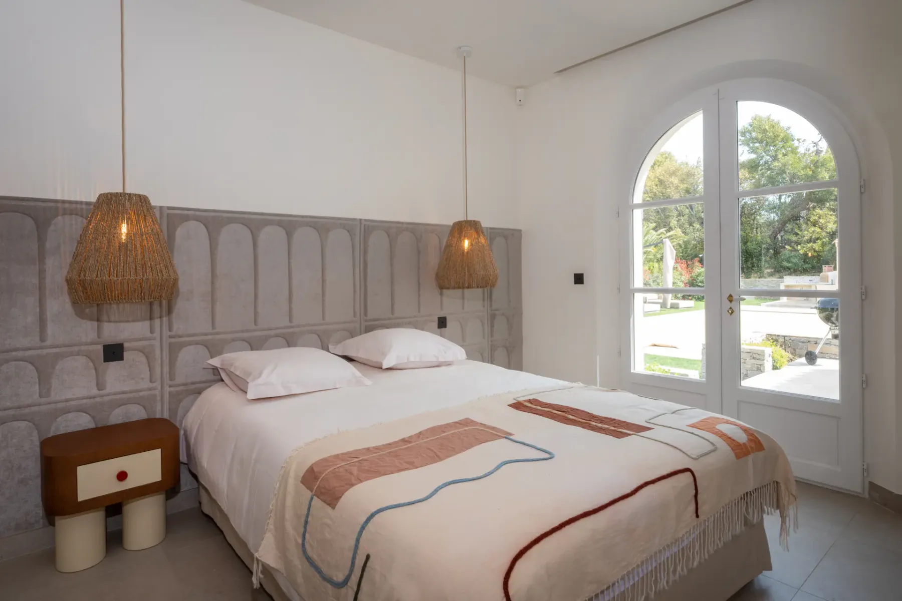 Dormitorio con cama doble, lámparas colgantes junto a la cama, cabecero revestido de azulejos y una puerta doble que da a una terraza exterior.