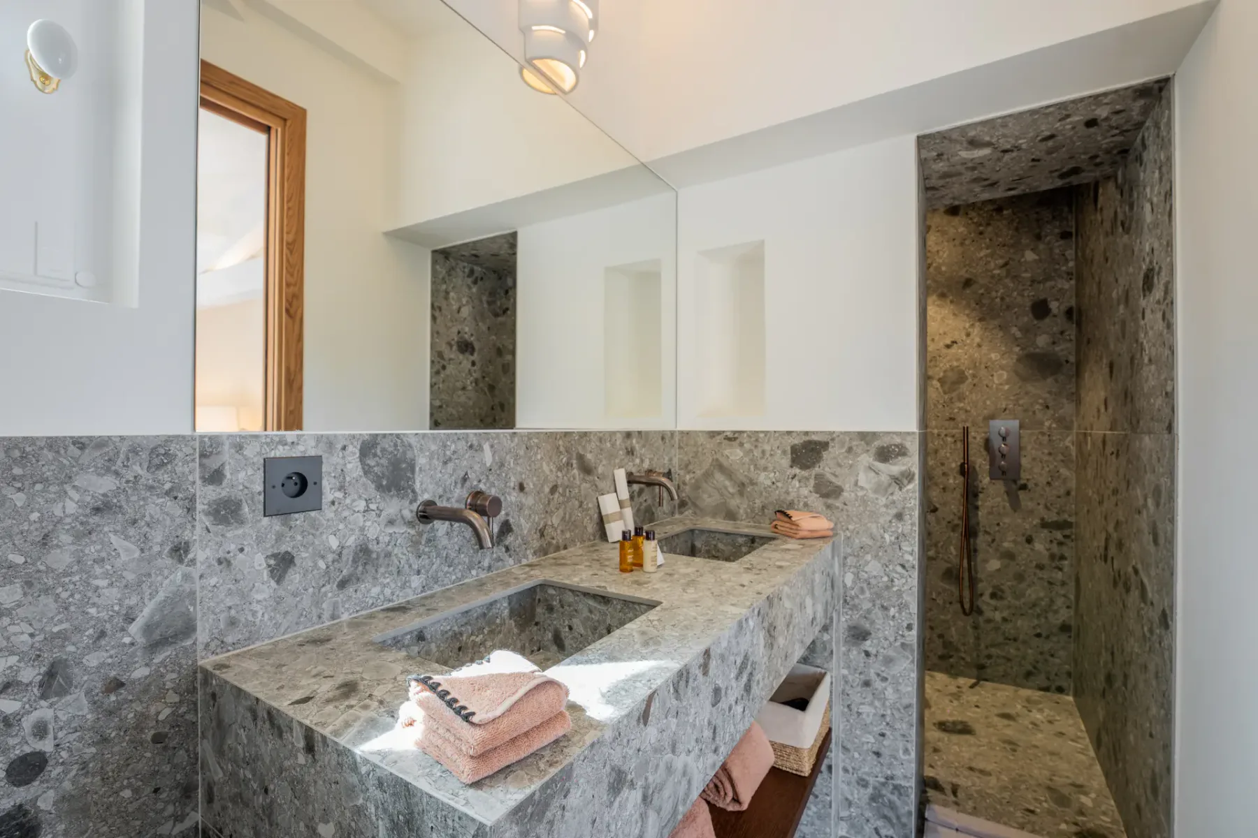 Baño moderno revestido con baldosas de piedra, con dos lavabos, un espejo y acceso a la ducha