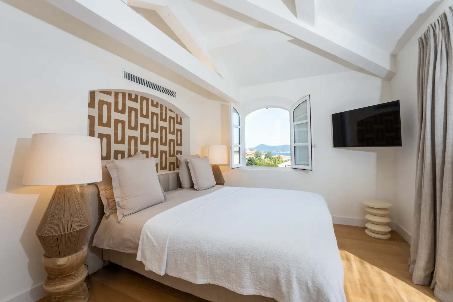 Dormitorio con una cama, un panel decorativo estampado y una ventana abierta con vistas al paisaje.