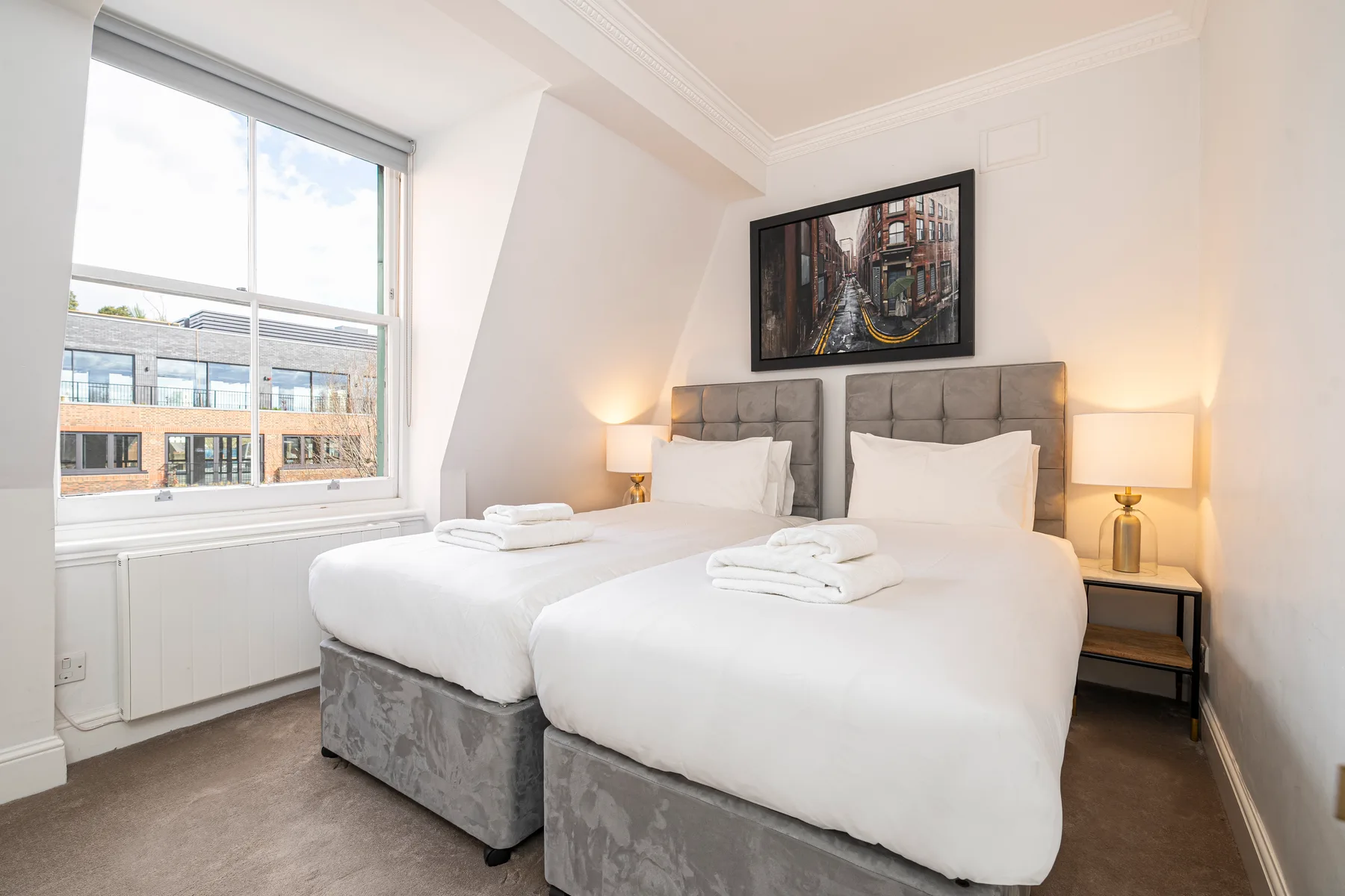 Un dormitorio luminoso tiene dos camas con ropa blanca junto a una ventana grande.