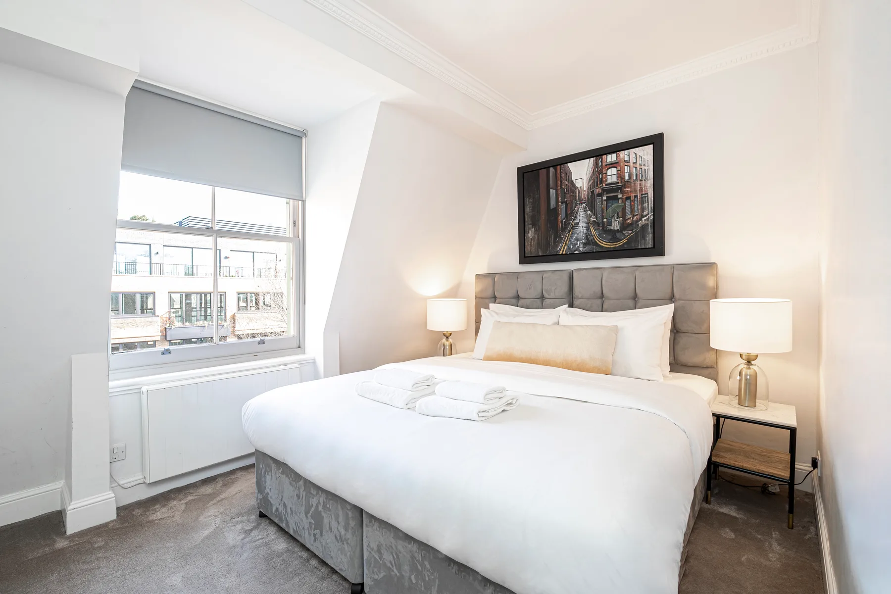 Un dormitorio con una cama grande, mesitas de noche y una ventana con vista a edificios cercanos.