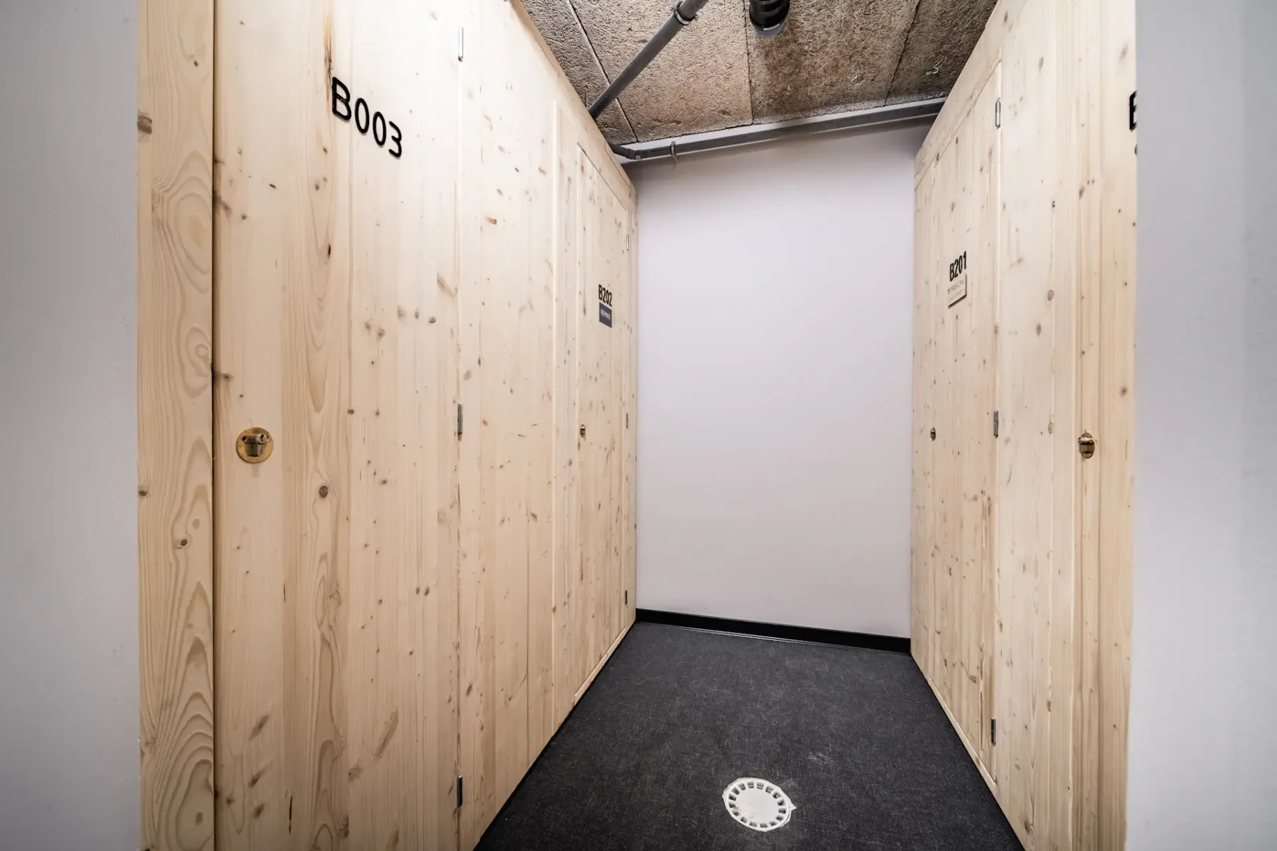 Wood-paneled storage unit corridor with three visible doors, labeled B003, under a ceiling with exposed pipes.
