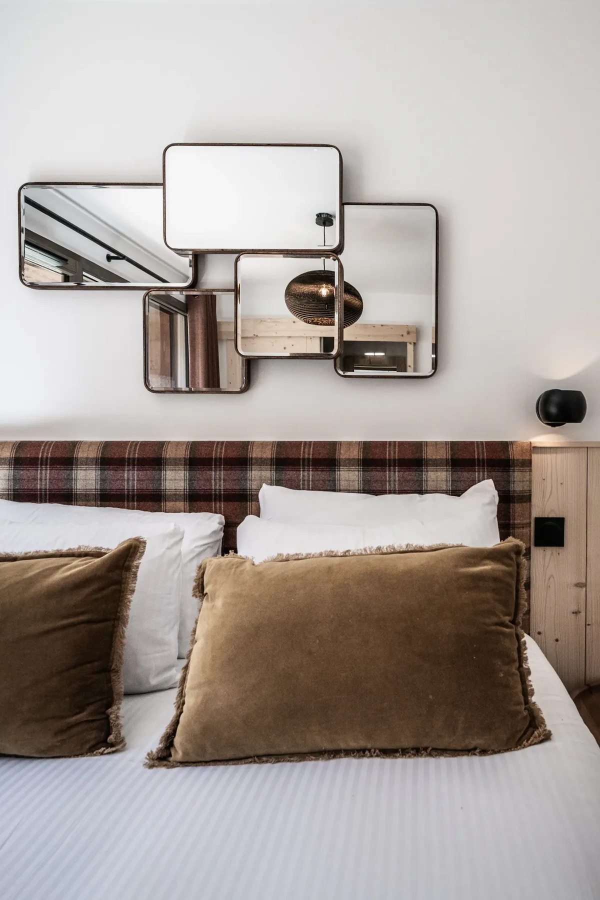 Bed with plaid headboard, brown pillows, white linens, and geometric mirrors above, in a cozy bedroom.