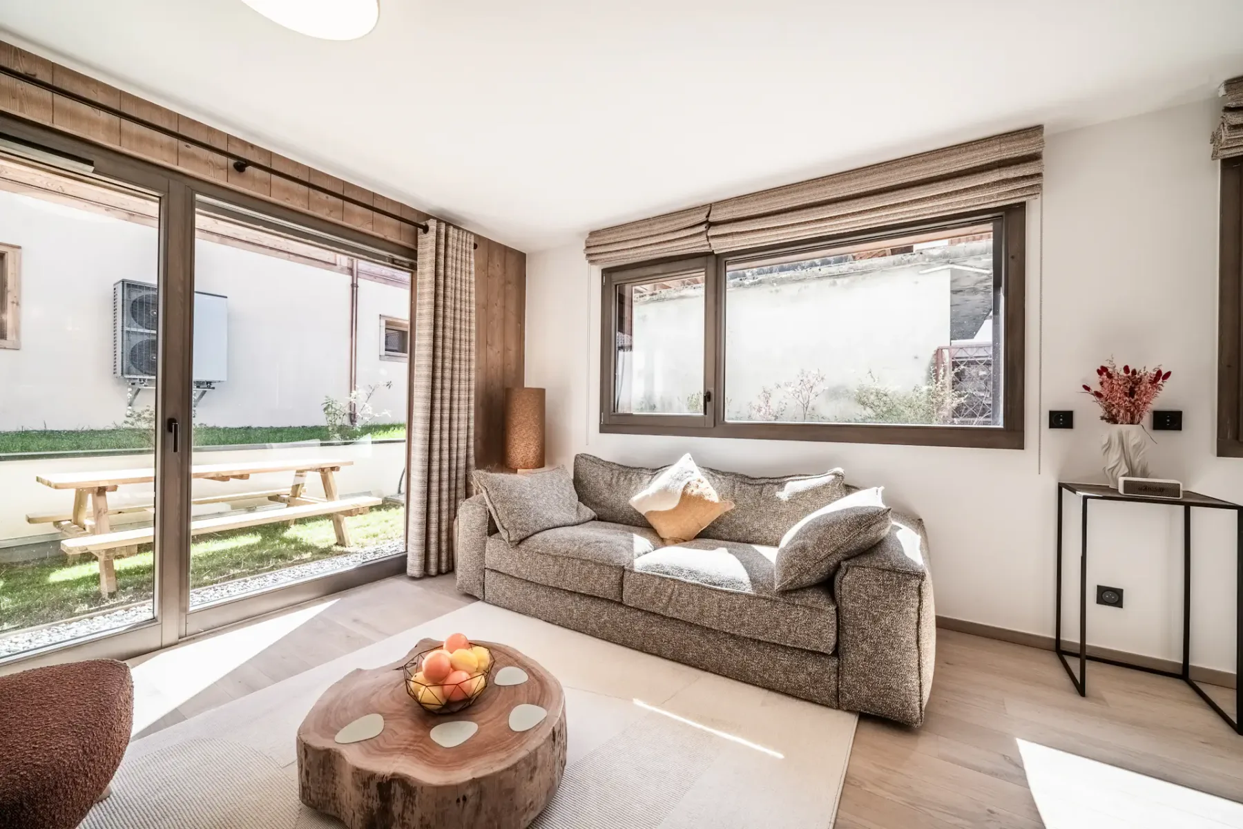 Una sala luminosa con un sofá gris junto a grandes ventanas con vistas a una zona de estar al aire libre.
