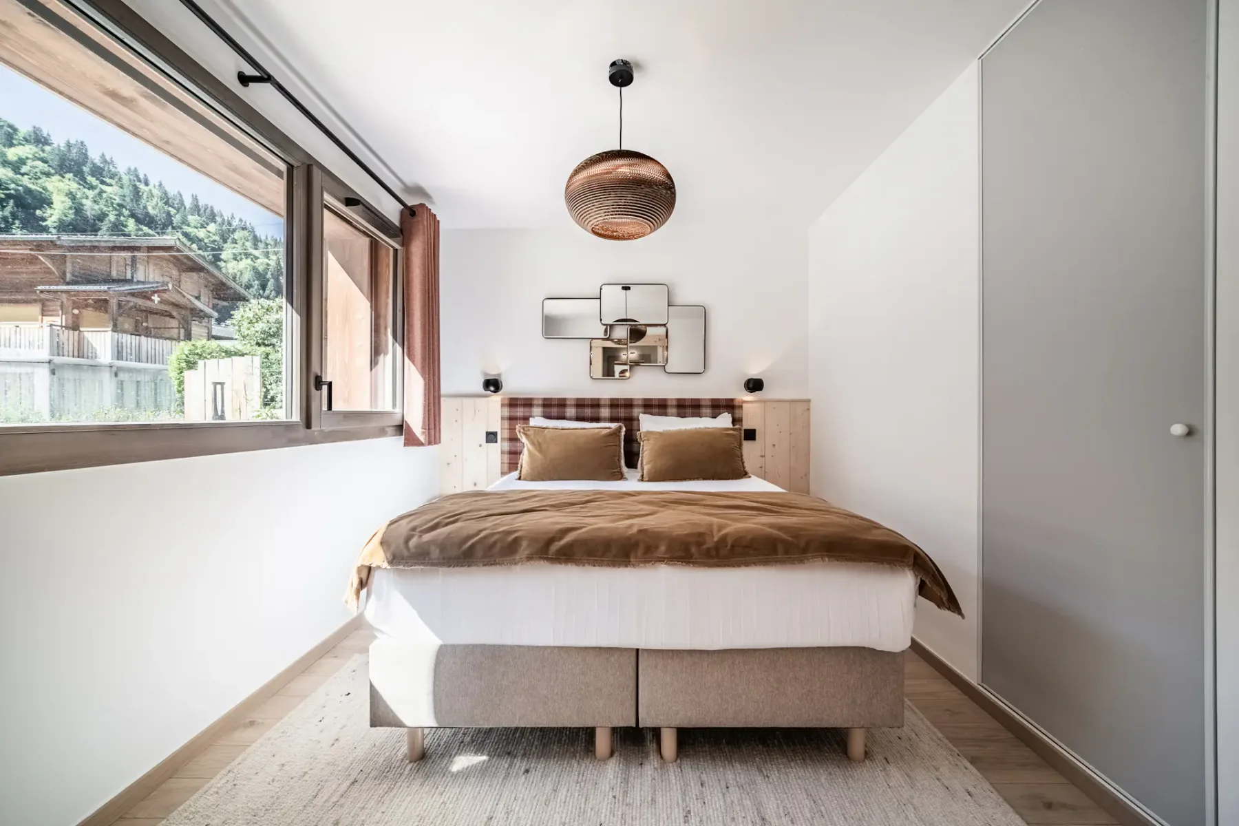 Cozy bedroom with large window, double bed, brown blankets, mirrored wall art, pink curtains, wooden accents, and a rustic lamp.