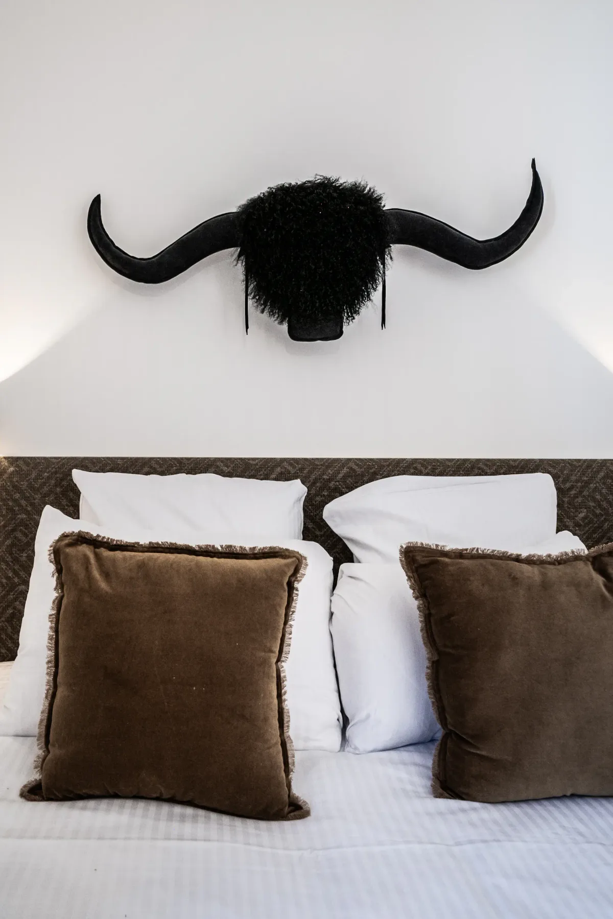 A cozy bed with white pillows and brown cushions, topped by a decorative bull head with long black horns on the wall above.