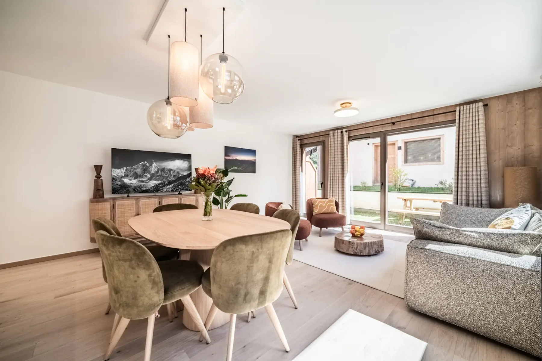 Bright, modern living room with round dining table, green chairs, pendant lights, and large windows overlooking a patio and garden.