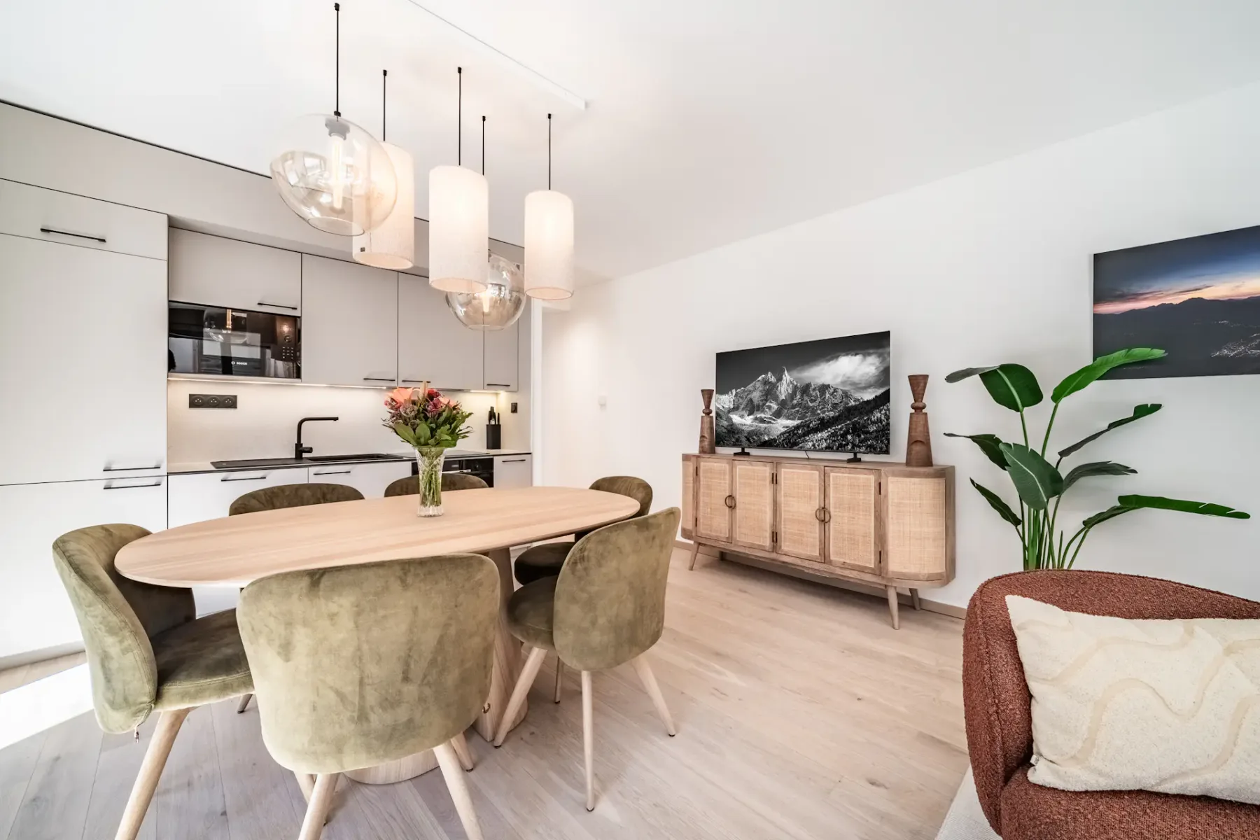 Modern open-plan kitchen and living area with white cabinets, a wood dining table, green chairs, and pendant lights.