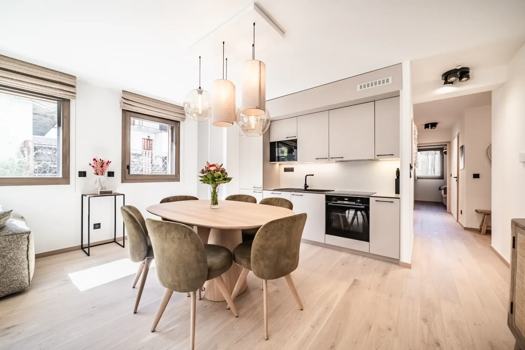 Modern open-plan kitchen and living area with white cabinets, a wood dining table, green chairs, and pendant lights.