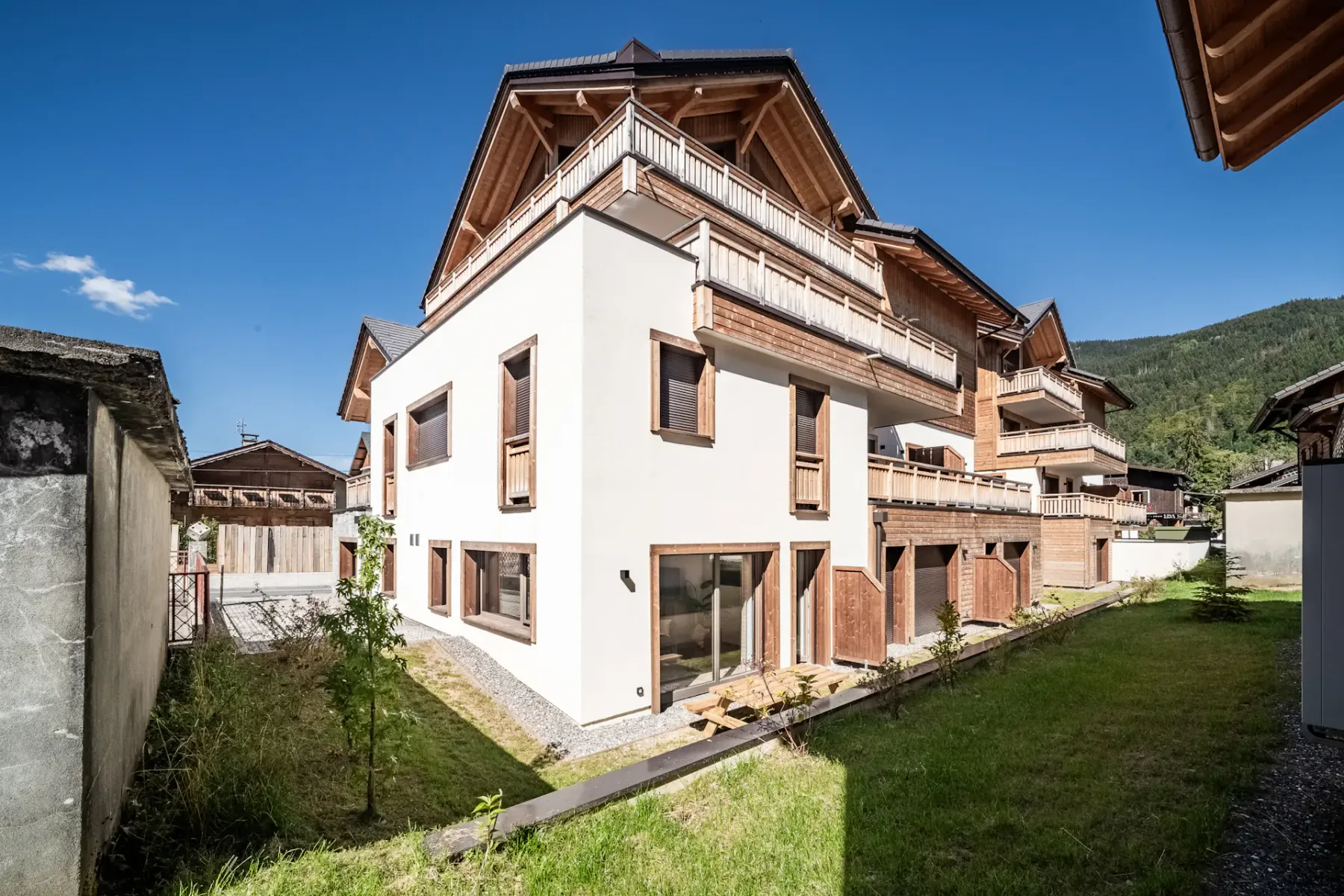Modern multi-story building with wooden accents and balconies, set in a grassy yard against a backdrop of mountains and a clear blue sky.