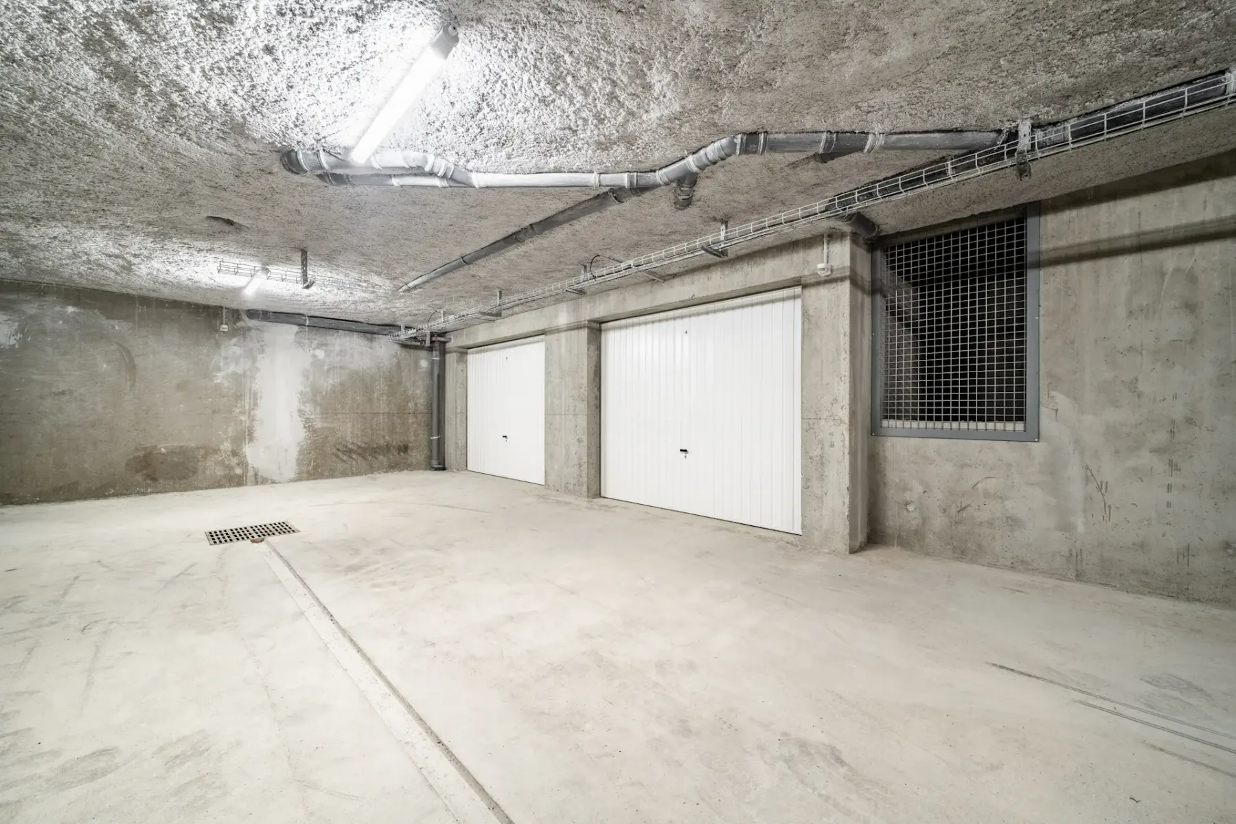 Empty underground parking garage with concrete walls, ceiling, and floor, featuring two closed white garage doors and fluorescent lighting.