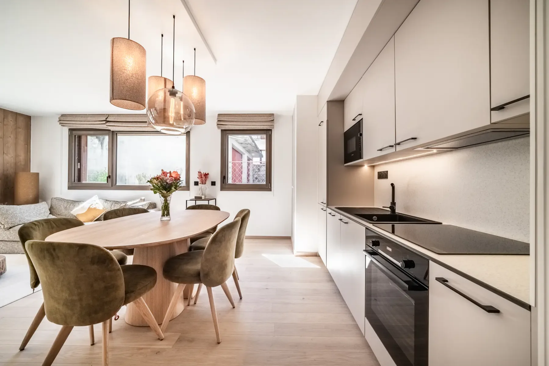 Modern open-plan kitchen and living area with white cabinets, a wood dining table, green chairs, and pendant lights.