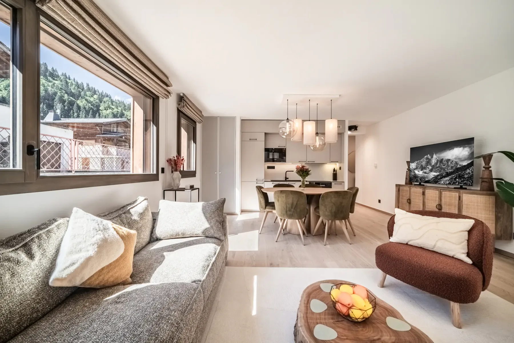 Sala de estar luminosa y moderna con mesa de comedor redonda, sillas verdes, lámparas colgantes y grandes ventanas con vistas a un patio y un jardín.