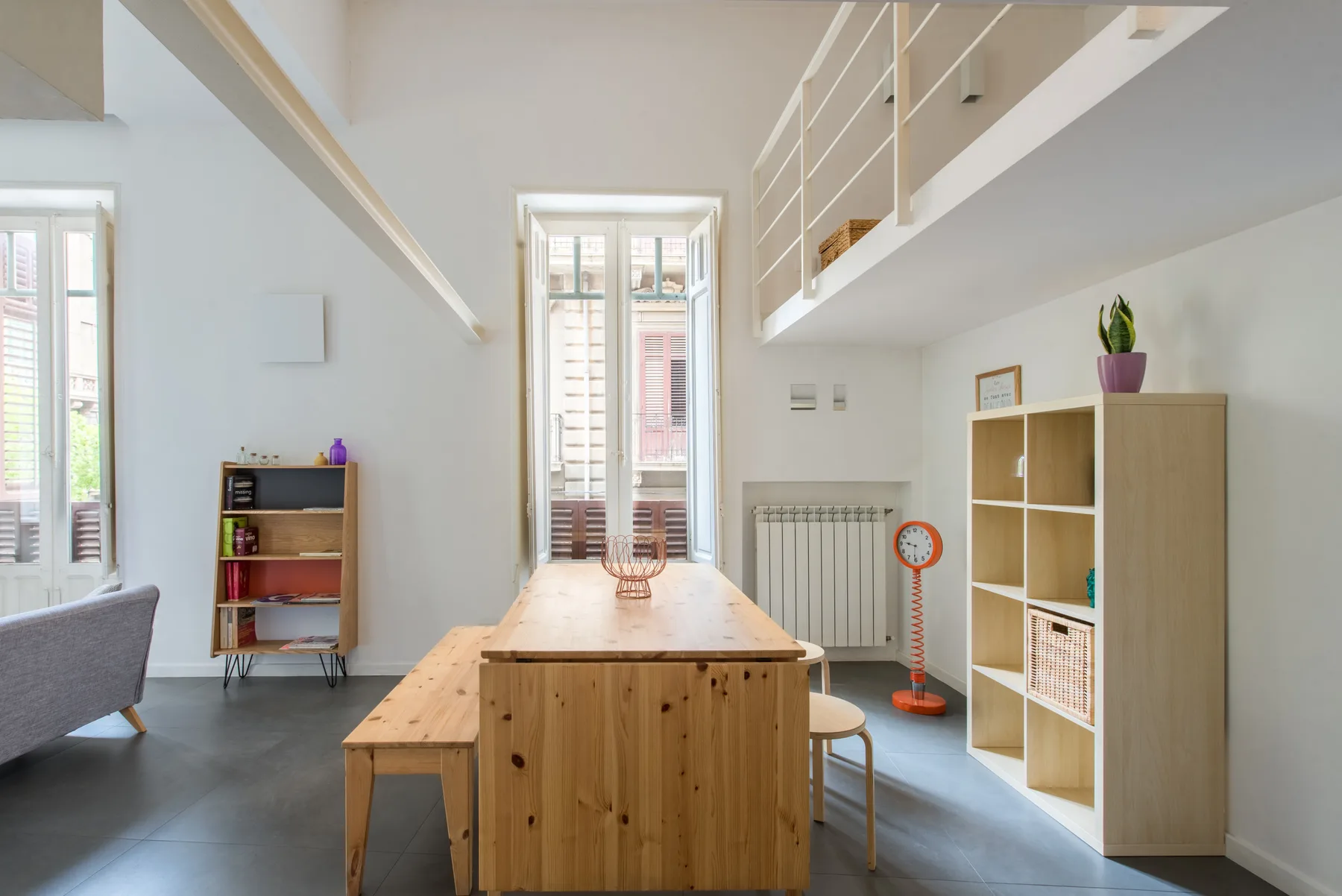 A bright loft room with a wooden dining table beneath a mezzanine.