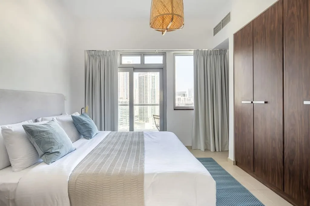 Un dormitorio luminoso con una cama bien hecha junto a grandes ventanas con cortinas.