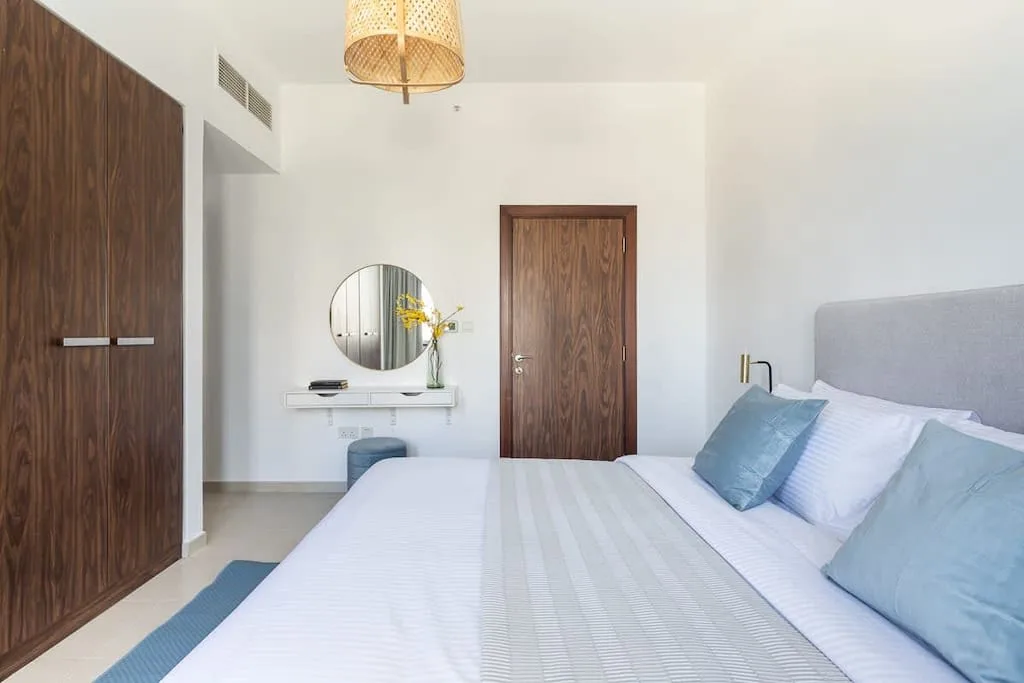 Un dormitorio luminoso con una cama tendida, una mesa auxiliar y un espejo redondo.