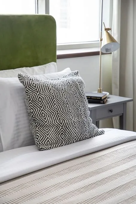 Un dormitorio con una cama, un cojín estampado y una mesita junto a la ventana.