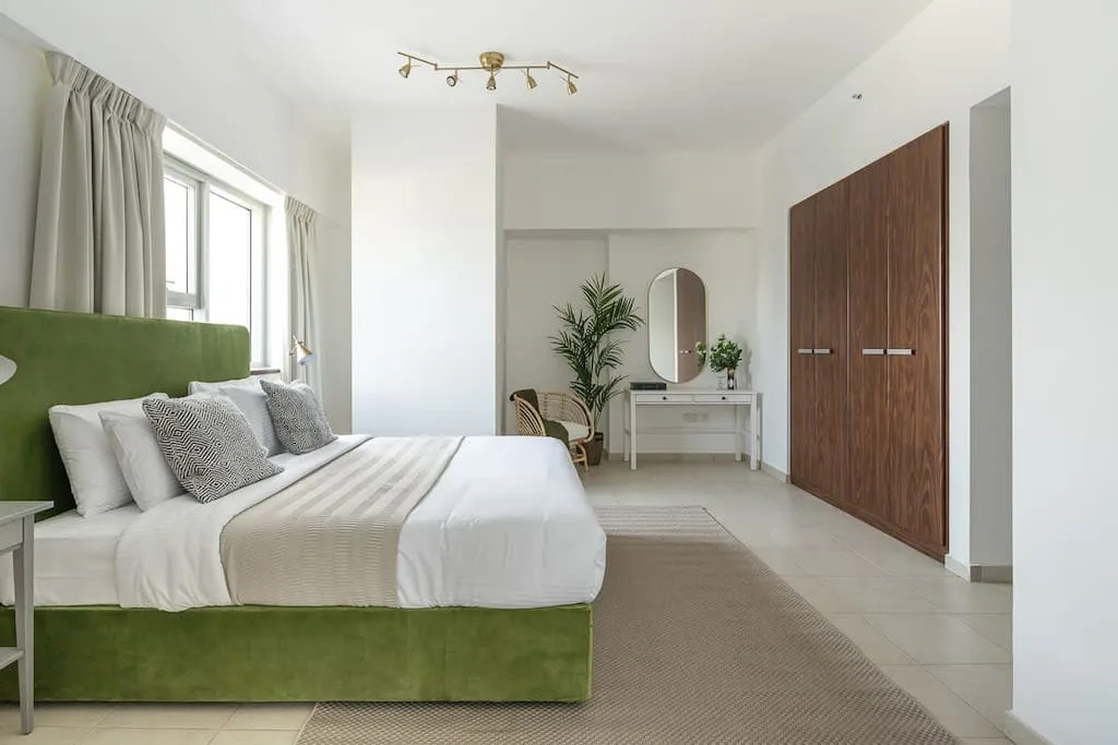 Un dormitorio luminoso con una cama verde, ventanas grandes y un tocador.