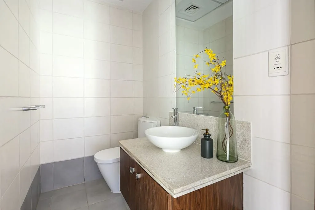 Un baño luminoso con azulejos, lavabo sobre encimera y una rama con flores amarillas.