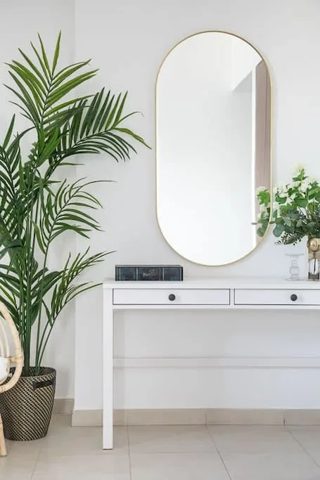 Un recibidor luminoso tiene una mesa consola blanca, un espejo ovalado y plantas frondosas.