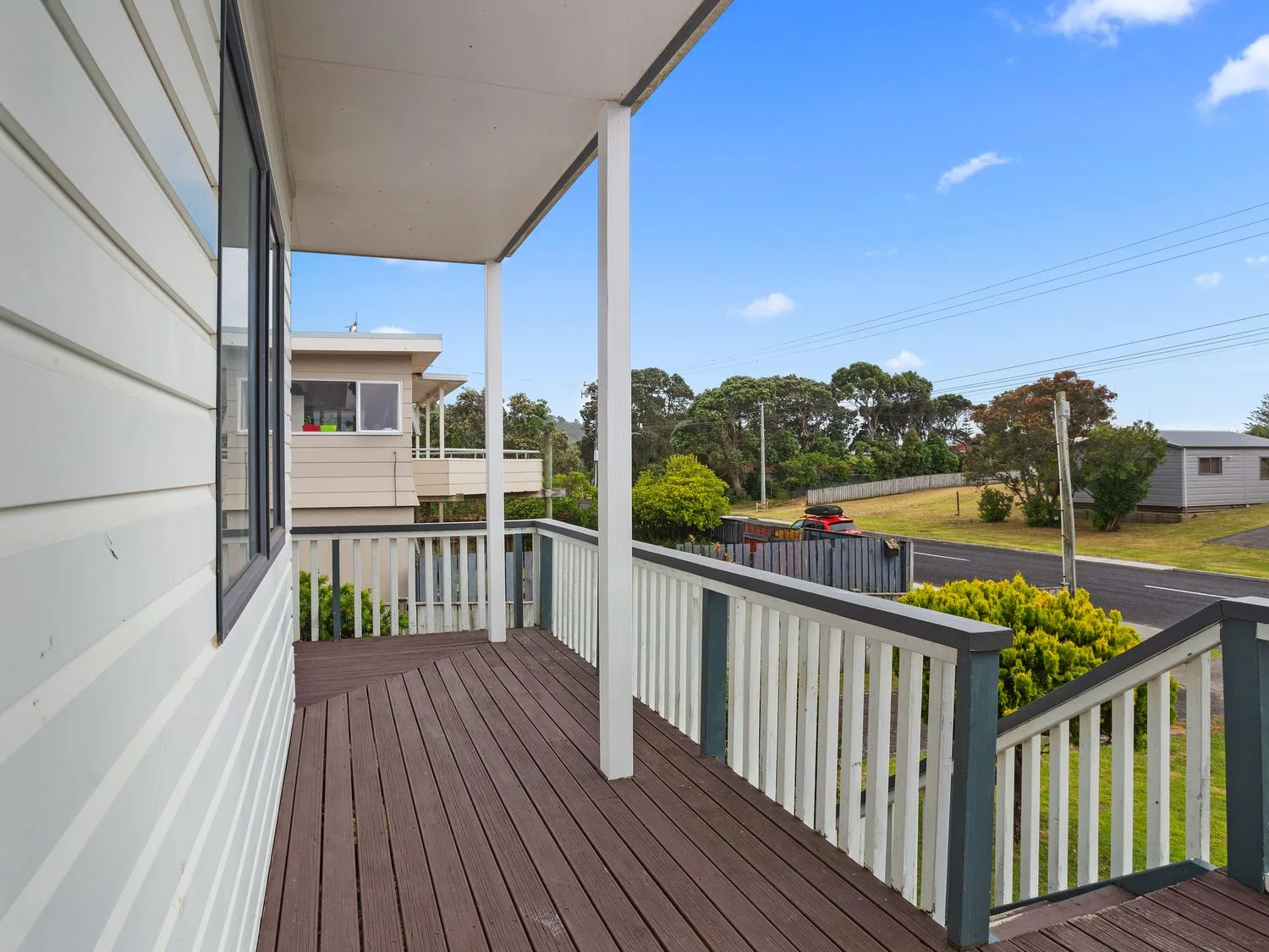 Un balcón cubierto de madera con barandillas blancas y vistas a una calle y a las casas vecinas en A Wave From It All - Waihi Beach