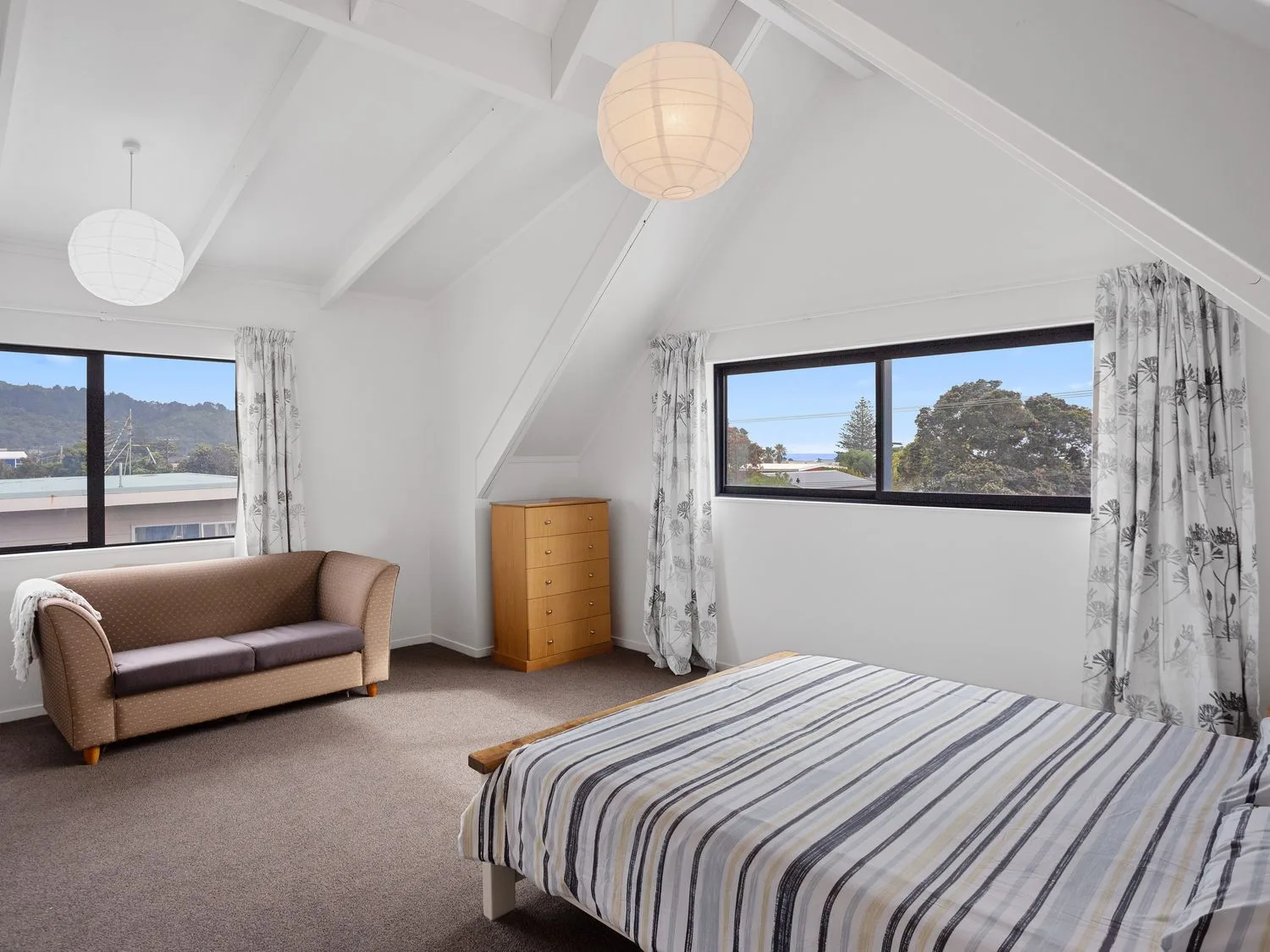 Un dormitorio con una cama, un sofá, una cómoda y ventanas con cortinas en A Wave From It All Waihi Beach