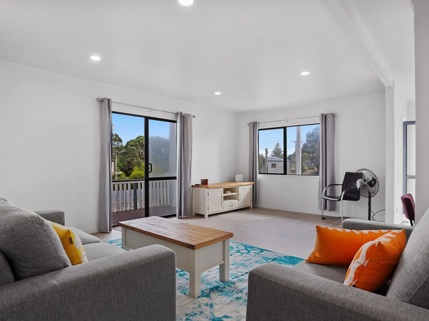 Una sala de estar con dos sofás grises, cojines naranjas y amarillos, una mesa de centro de madera, un armario blanco, una silla y un ventilador de pie en A Wave From It All - Waihi Beach