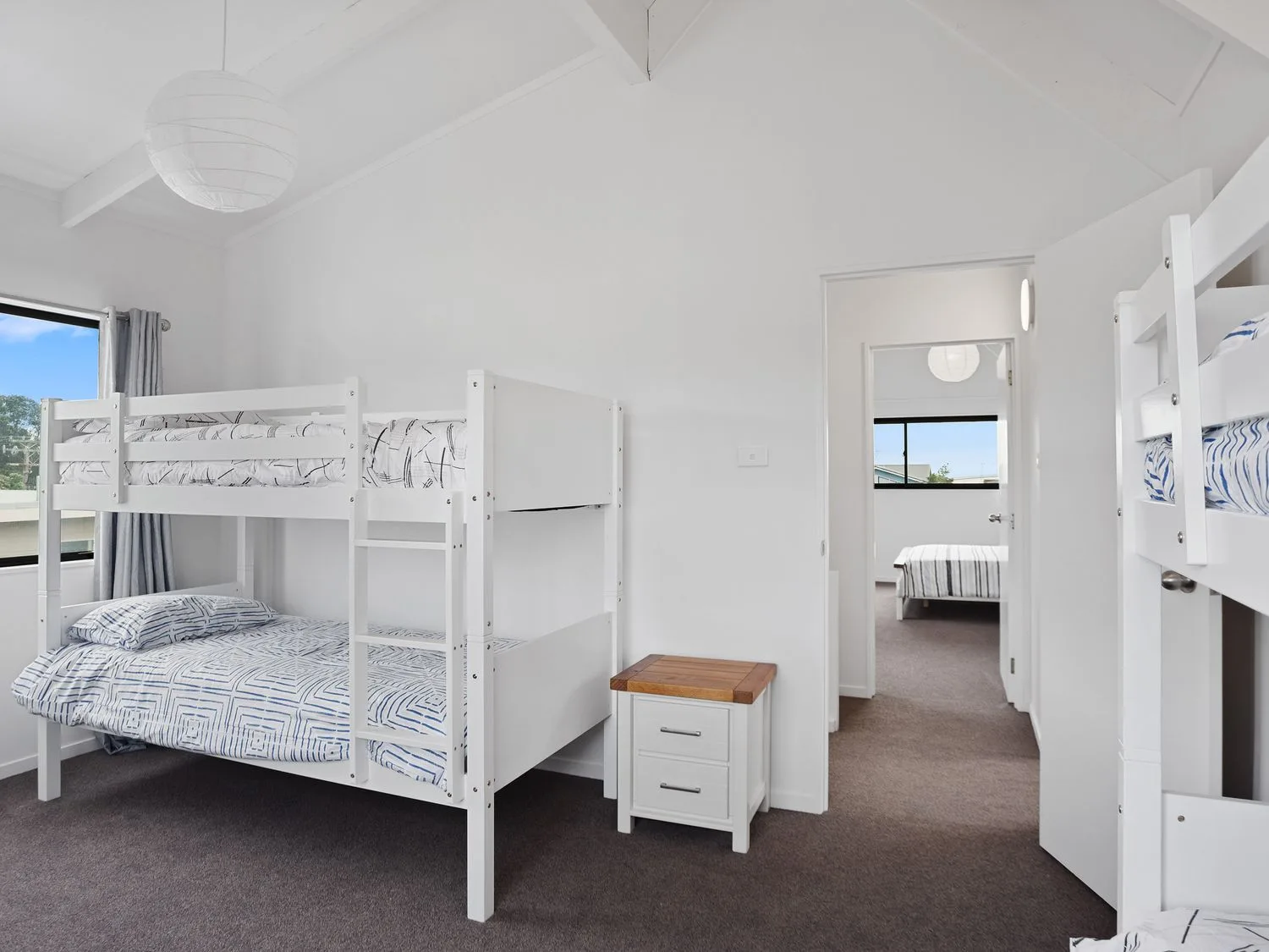 Un dormitorio con dos literas blancas, una pequeña mesita de noche blanca y vistas a otro dormitorio en A Wave From It All - Waihi Beach