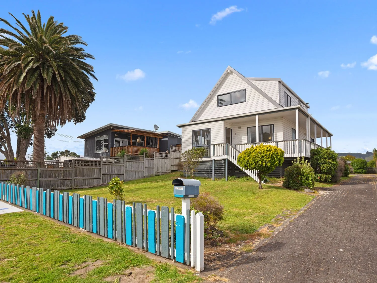 Una casa blanca con un porche que la rodea, junto a una palmera y una cerca de madera pintada en A Wave From It All Waihi Beach