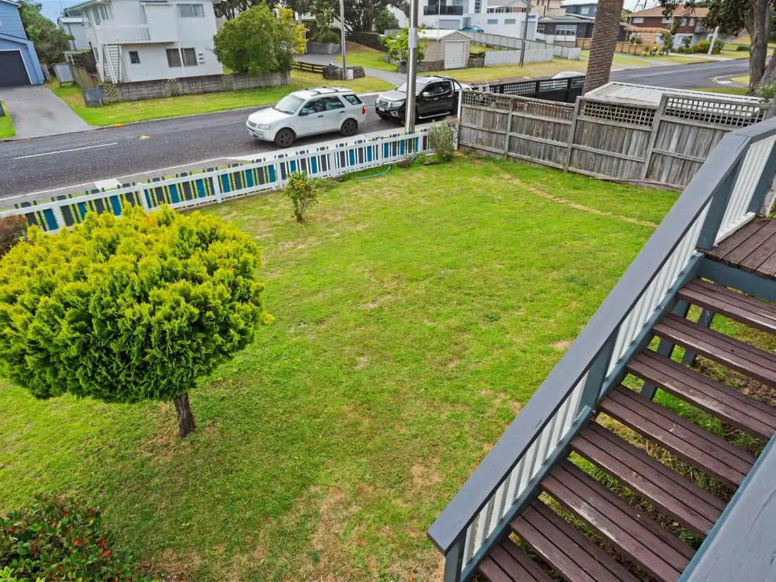 Un jardín delantero con césped, un árbol pequeño y una cerca blanca y azul, junto a una calle con coches aparcados en un barrio residencial en A Wave From It All - Waihi Beach