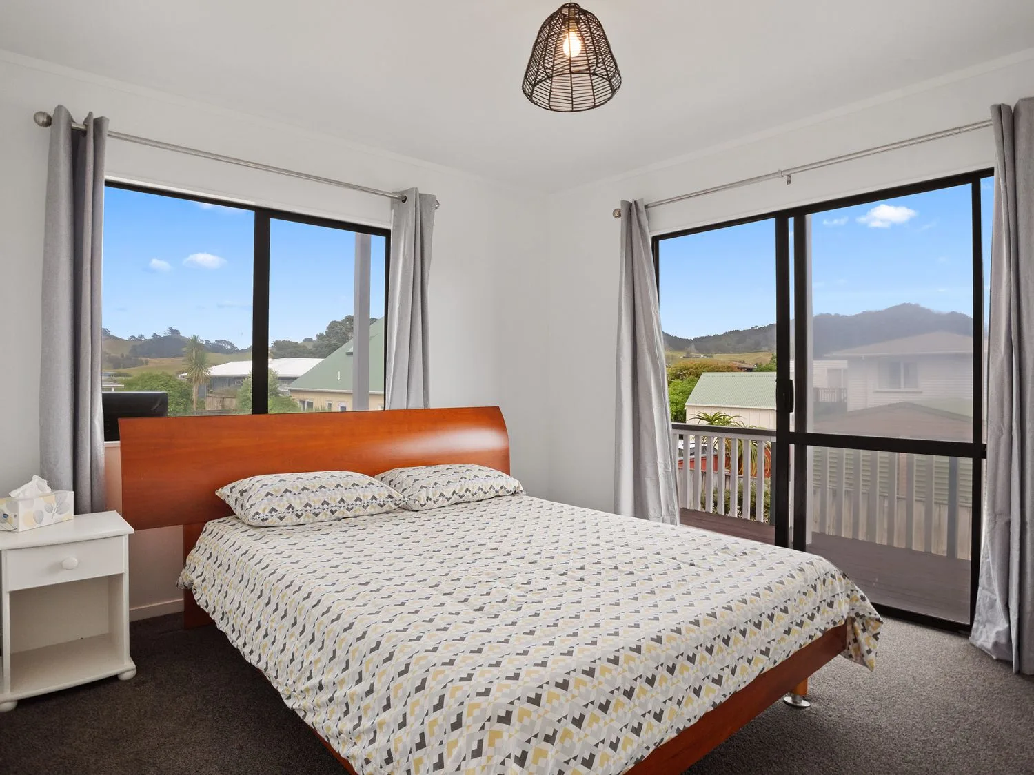 Un dormitorio con una cama cubierta con ropa de cama estampada, junto a dos grandes ventanas con cortinas grises en A Wave From It All - Waihi Beach Holiday Home