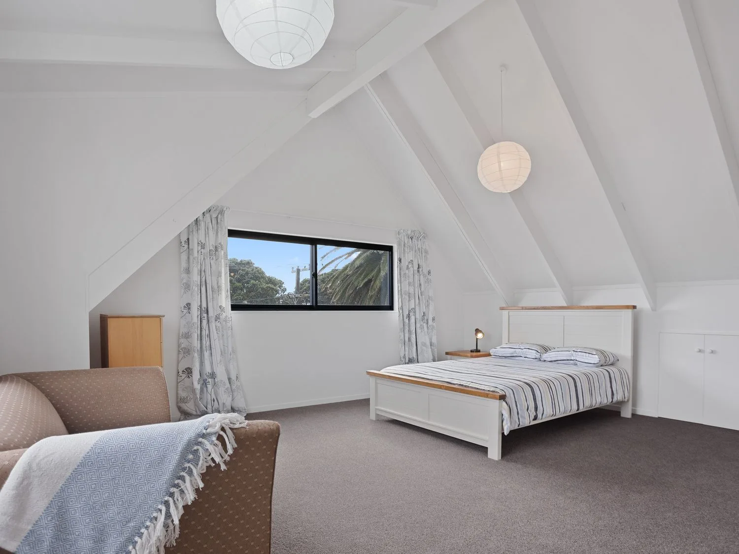 Un dormitorio con una cama a rayas, una mesita de noche con una lámpara, una ventana con cortinas y una silla marrón con una manta en A Wave From It All Waihi Beach