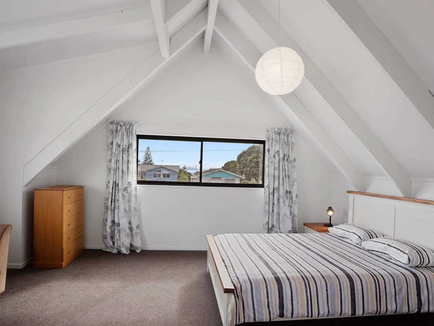 Un dormitorio con una cama, ropa de cama a rayas, una cómoda de madera y una ventana con cortinas en A Wave From It All - Waihi Beach Holiday Home, en Waihi Beach