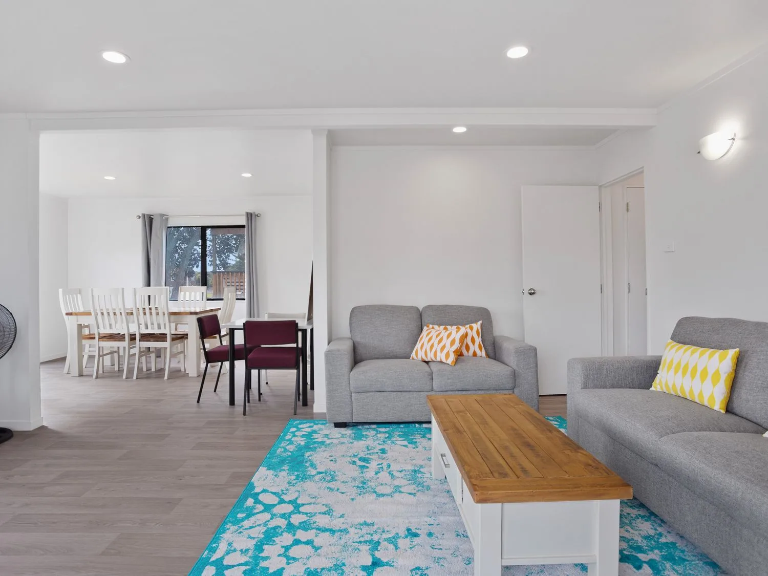 Una sala de estar con asientos grises, una mesa de centro de madera y una alfombra azul estampada junto a un comedor.