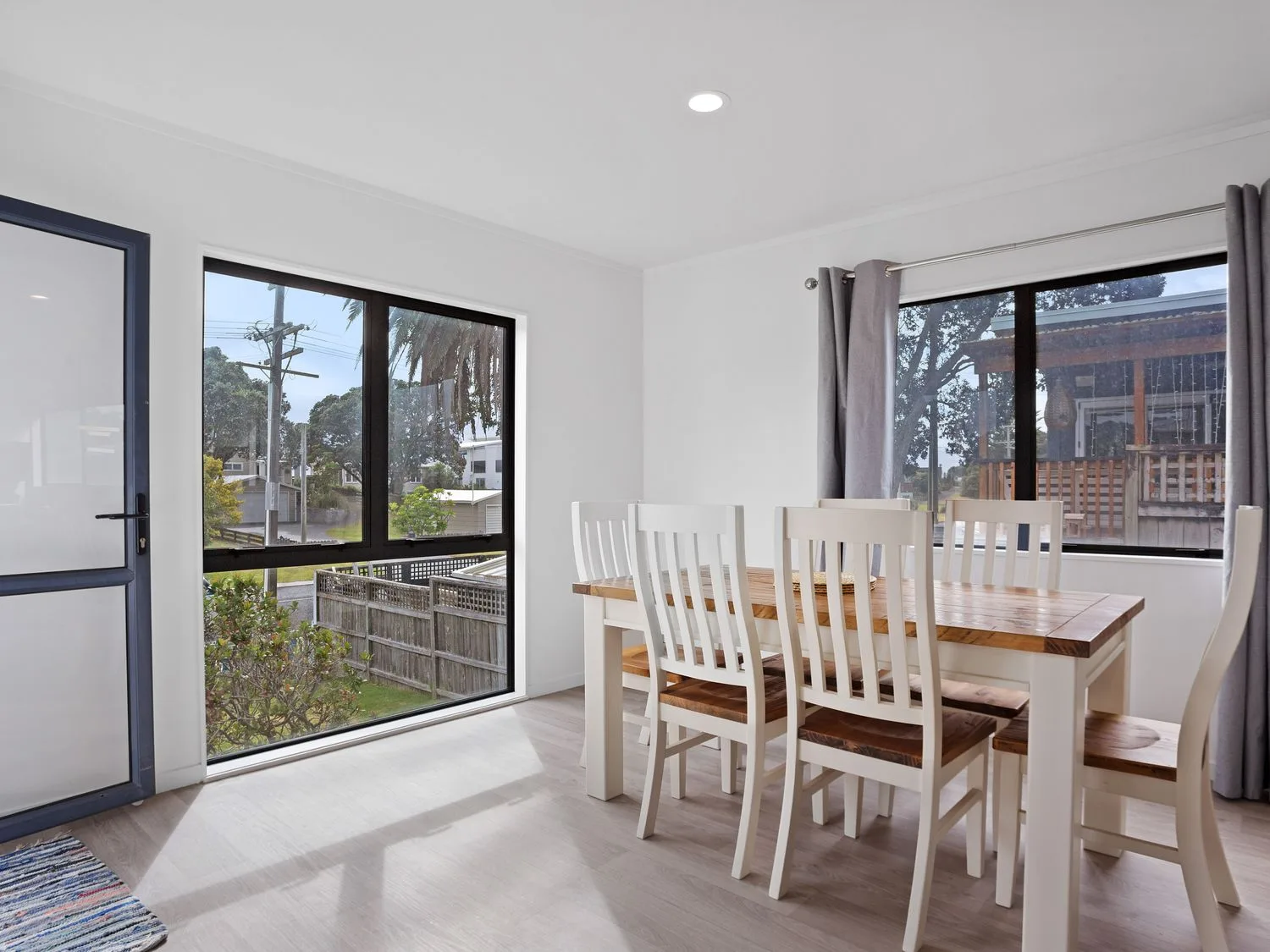 A dining room with a wooden table and six white chairs near large windows with gray curtains at A Wave From It All - Waihi Beach Holiday Home in Waihi Beach