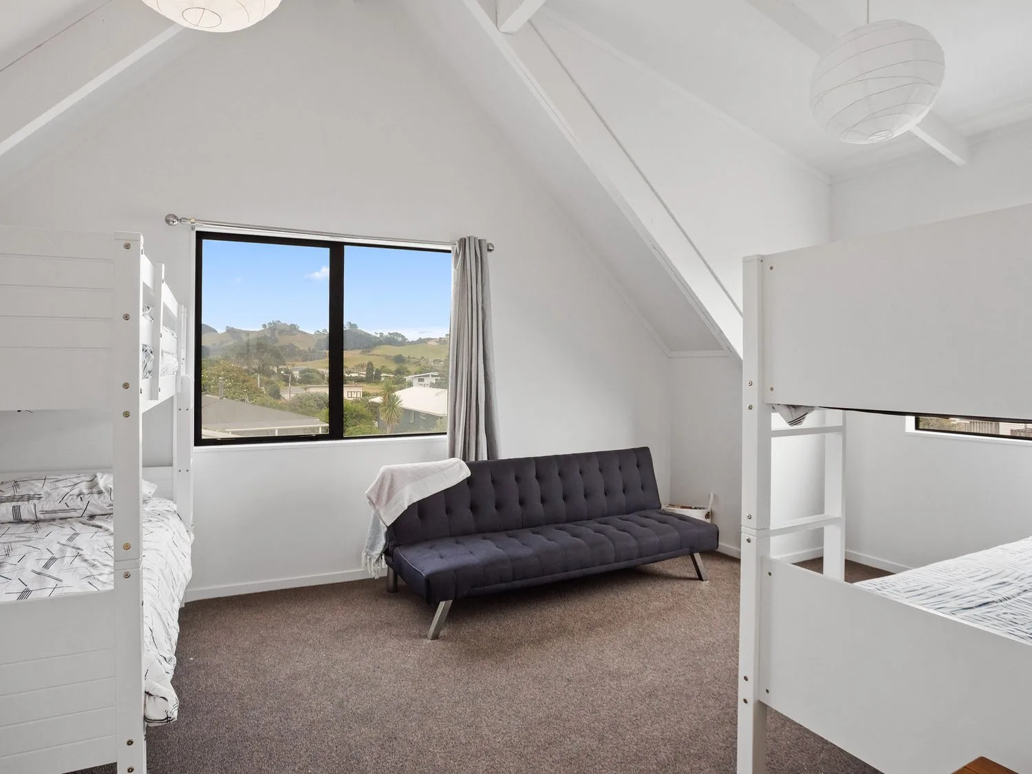 Un dormitorio con dos literas blancas, un sofá negro y una ventana desde la que se ve un paisaje en A Wave From It All - Waihi Beach Holiday Home, en Waihi Beach