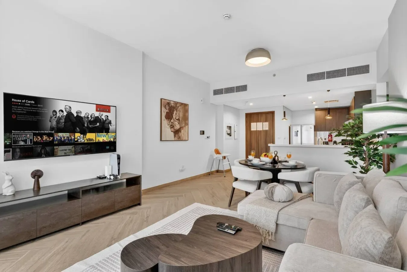 A bright living room with seating, a television, and an open kitchen in the background.