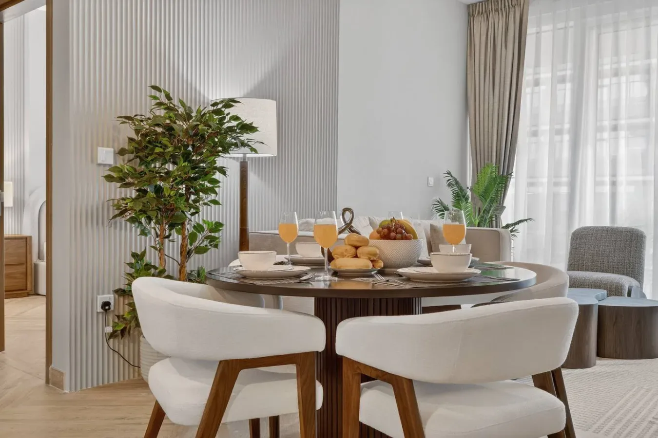 A dining area with a round table set with dishes and fruit beside a bright window.