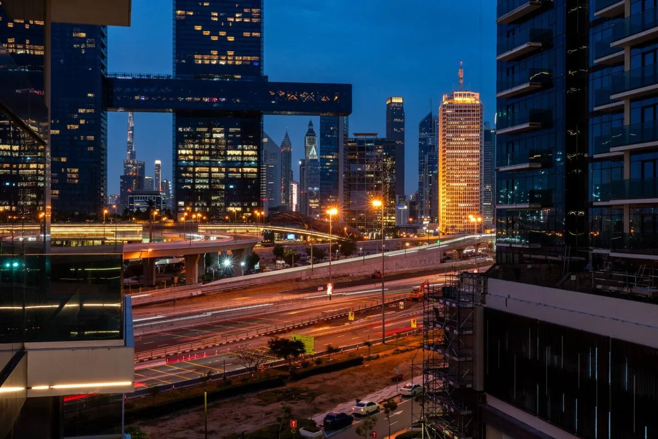 A city skyline at dusk rises above illuminated roads and elevated tracks.