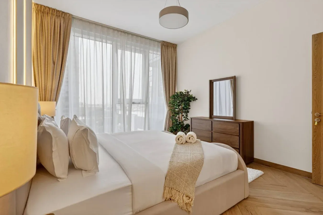 A bright bedroom with a neatly made bed beside a large curtained window.