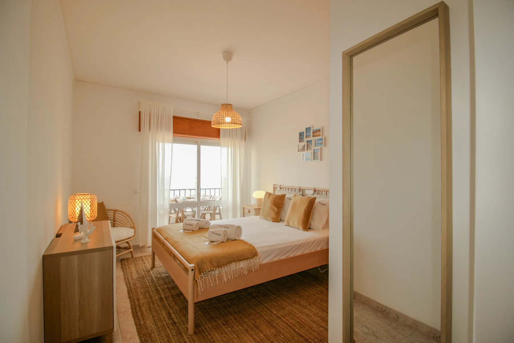 A bright bedroom with a wooden bed, bedside cabinet, and window with sheer curtains.
