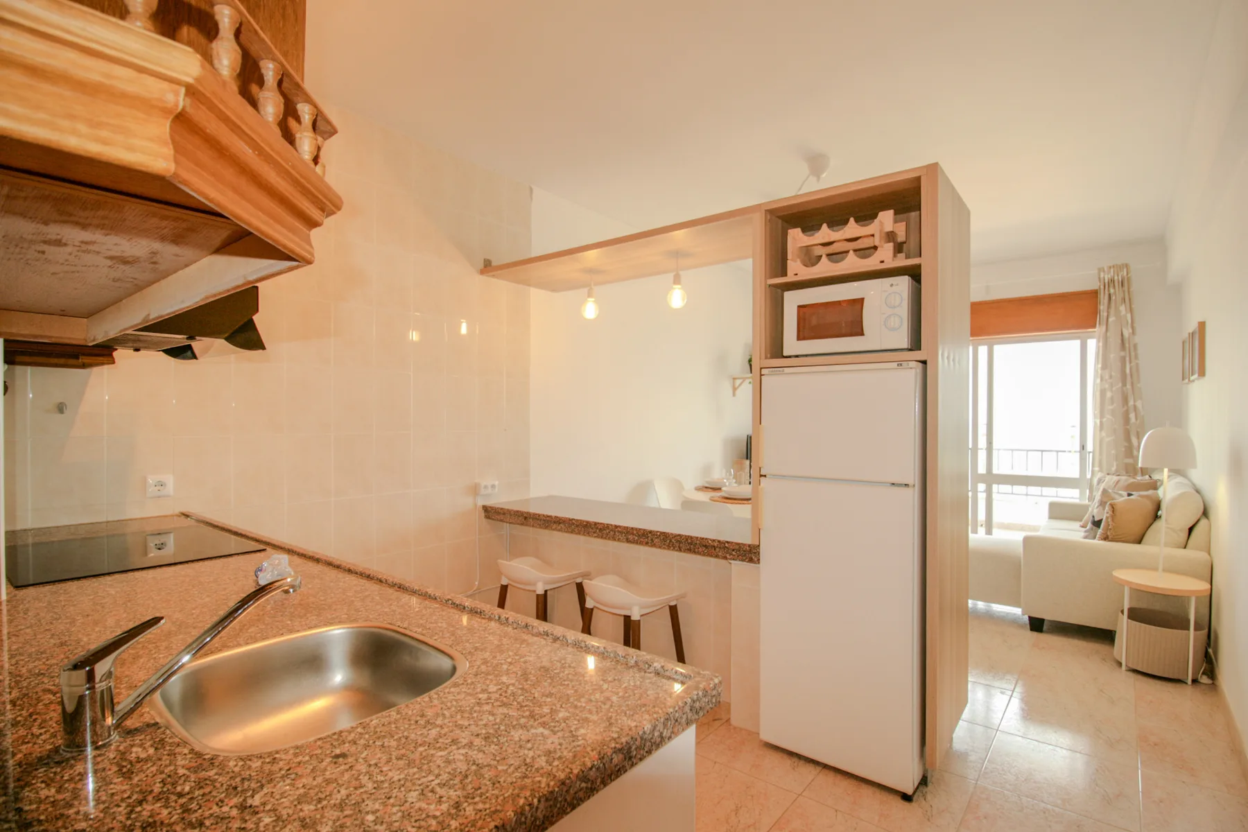 Una cocina con encimera y fregadero se abre a una sala de estar junto a una ventana luminosa.
